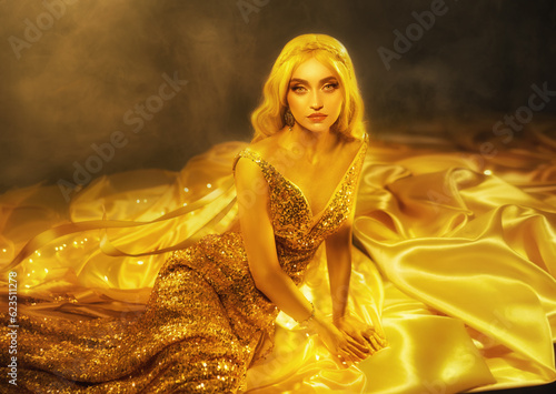 fantasy woman blonde in long golden evening shine glow dress lies in satin fabric on floor, beauty face looks at camera. Art photo portrait fairy girl princess. gold plated paint skin creative makeup