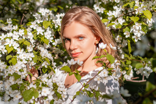 Portrait of a girl in a white dress in blooming apple trees. The girl poses against the background of flowers in the spring park.