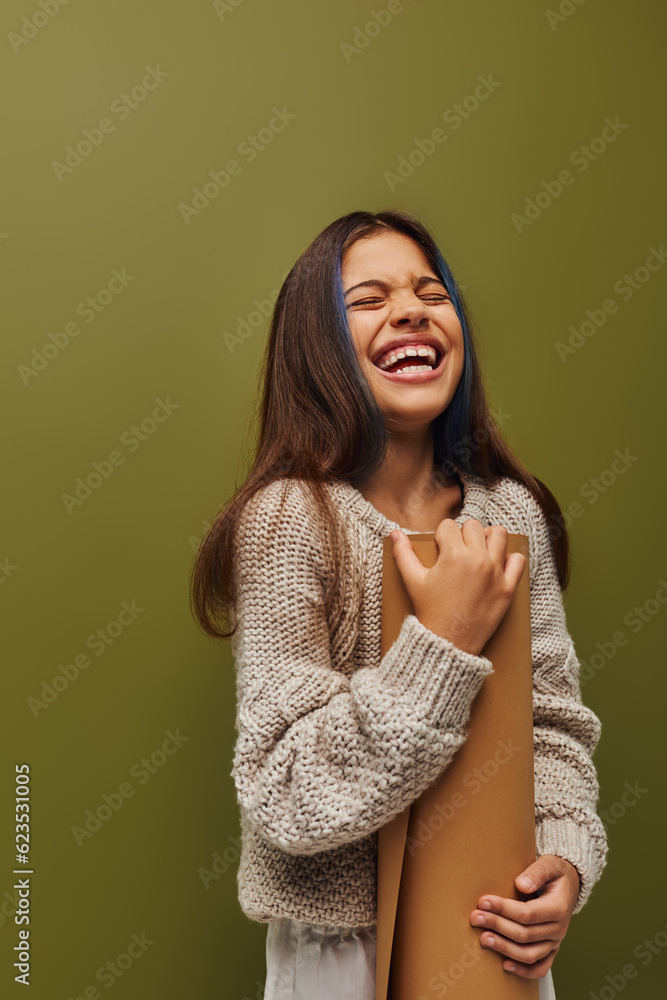 Cheerful and stylish preteen girl in modern knitted sweater and autumn outfit holding rolled paper and laughing while standing isolated on green, girl radiating autumn vibes concept Cheerful and stylish preteen girl in modern knitted sweater and autumn outfit holding rolled paper and laughing while standing isolated on green, girl radiating autumn vibes concept