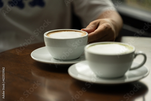 Closeup hand holding white cup of hot coffee latte and blurry hot green tea cup on wood background in restaurant.Best of menu in the coffee shop.Soft focus.