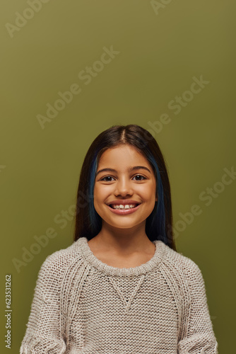 Portrait of positive and stylish brunette preteen girl with dyed hair wearing modern cozy knitted sweater while looking at camera while standing isolated on green, modern preteen fashion