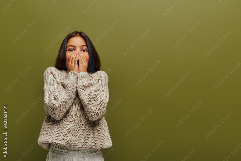 Scared preteen kid with dyed hair wearing stylish knitted and cozy sweater while covering face and looking at camera isolated on green, contemporary fashion for preteen concept Scared preteen kid with dyed hair wearing stylish knitted and cozy sweater while covering face and looking at camera isolated on green, contemporary fashion for preteen concept