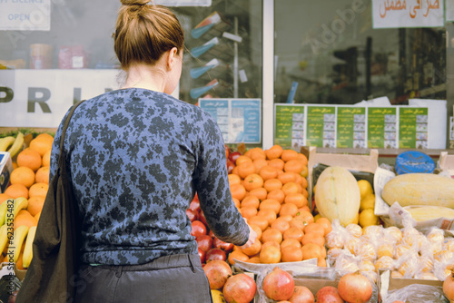 Rear view of single female young-adult buying food at a food store, choosing what to buy at the fruit stand outside, looking at food carrying a tote bag. Extended arm. Signs in English and Arabic.