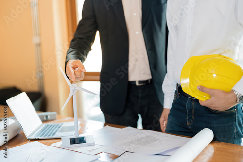 White energy wind turbine model on the desk Hands of two Asian male architects are pointing, holding helmets. And there is a blueprint about the design of the construction industry in the office.