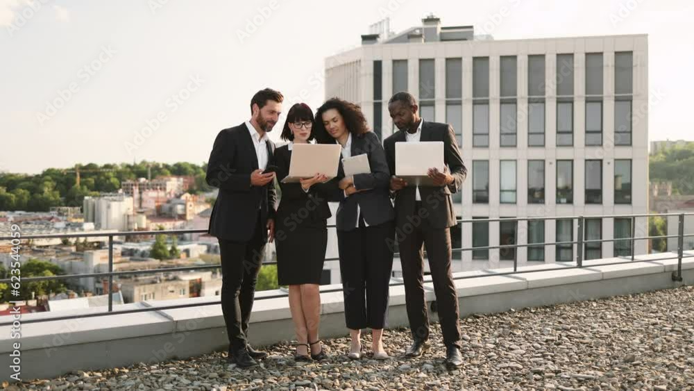 Diverse team of foreign investors standing on flat rooftop with personal gadgets and discussing marketing strategy of new business project. Officially dressed colleagues watching online presentation. Diverse team of foreign investors standing on flat rooftop with personal gadgets and discussing marketing strategy of new business project. Officially dressed colleagues watching online presentation.