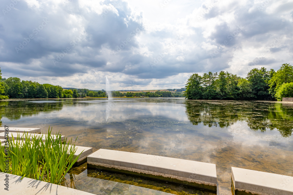 Echternach lake with concrete platforms on shores, waterjet gushing from fountain and green leafy trees in background, reflection on water surface, panoramic landscape, cloudy summer day in Luxembourg