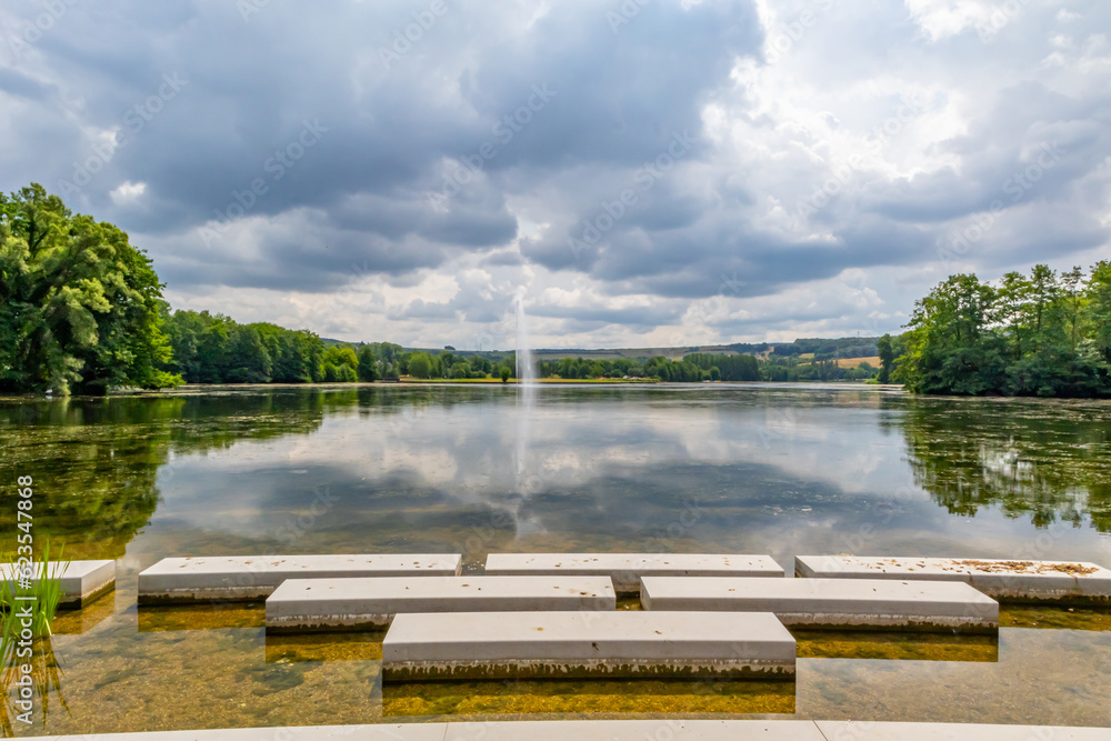Fototapeta premium Panoramic landscape of Lake Echternach with waterjet from a fountain, green leafy trees on background, concrete platforms on shore, reflection on water surface, cloudy summer day in Luxembourg