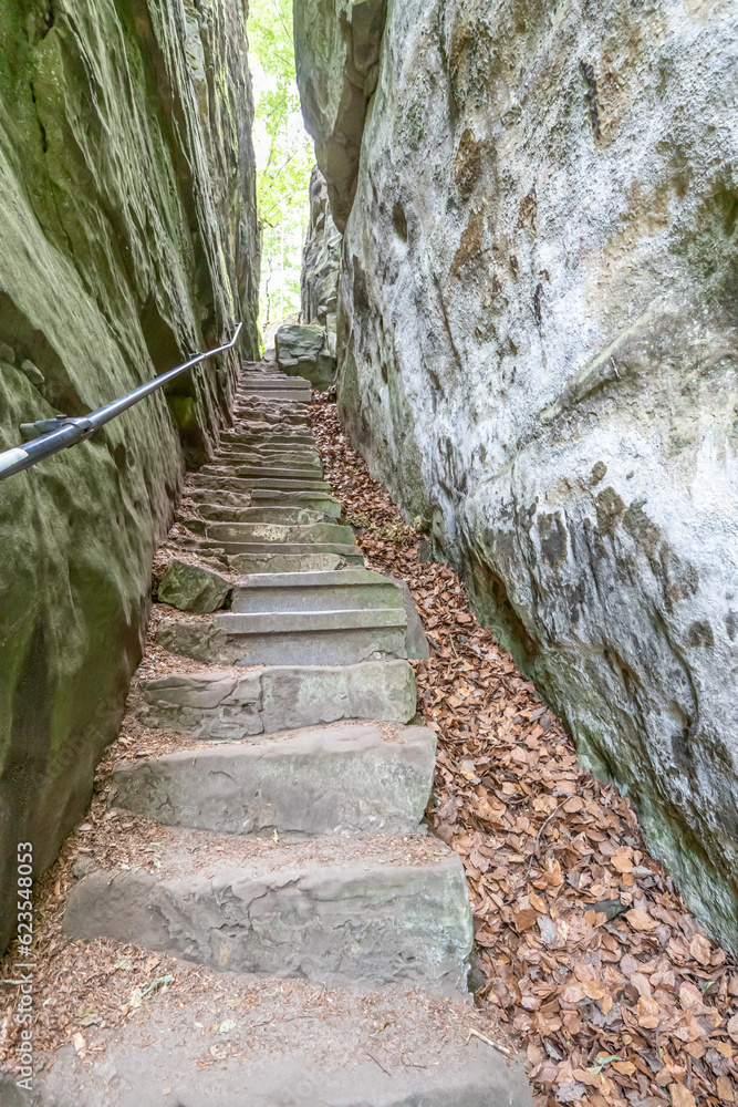 Narrow slot with a staircase between two rock formations in Teufelsschlucht nature reserve, sunlight in background, view from an upward perspective, sunny summer day in Ernzen, Germany