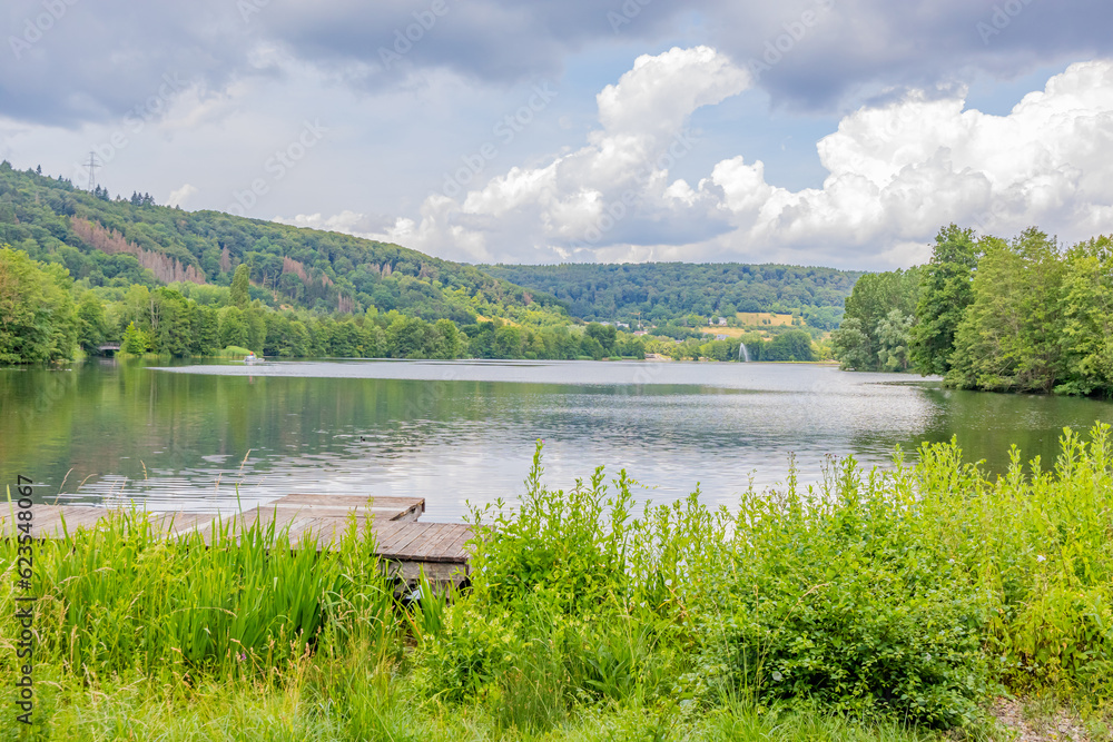 Lake Echternach landscape with fishing platform on shore, calm water, hills with abundant leafy trees on misty background, reflection on water, sunny summer day with abundant clouds in Luxembourg