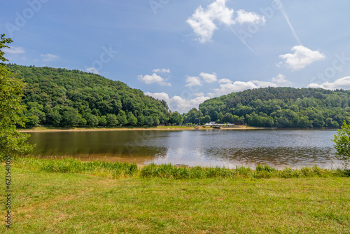 Landscape of Stausee Bitburg reservoir lake, calm water, mountains with abundant trees with green foliage in background, sunny spring day with a blue sky in Germany