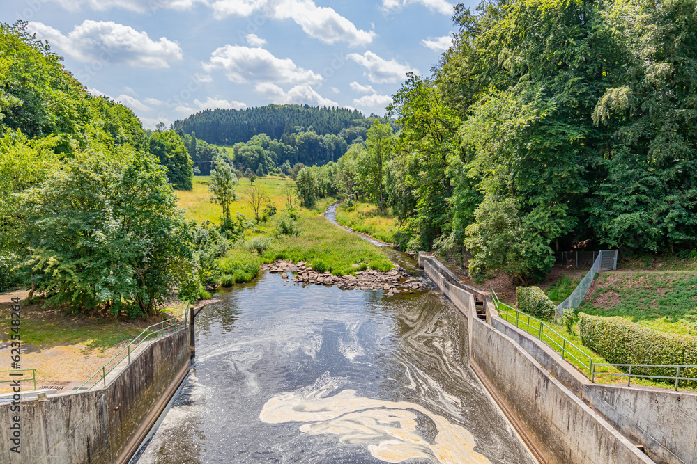 Water flowing from Staumauer Bitburger dam into Prum river, valley and mountains with leafy trees in background, brown spot on water surface, Stausee Bitburg reservoir, sunny spring day in Germany