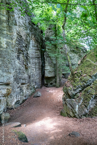 Hiking trail between huge rock formations in nature reserve Teufelsschlucht, walls with moss, uneven texture and grooves, trees with green foliage in background, sunny summer day in Ernzen, Germany