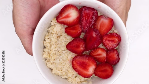 slice of strawberry on porridge, oats gruel top view flat lay. woman hand arranging fruits. cutting with knife strawberry in 2 parts,white plate 4k