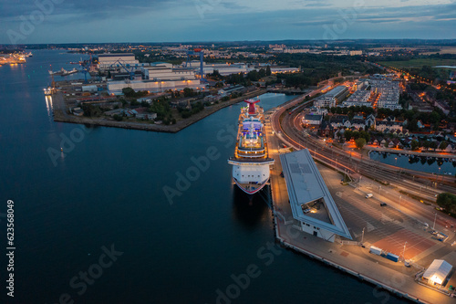Rostock, Warnemuende, Germany, aerial dusk view of illuminated cruise ship "Carnival Pride" of Carnival Cruise Line, moored at Cruise Ship Terminal, Warnemuende city in the background 