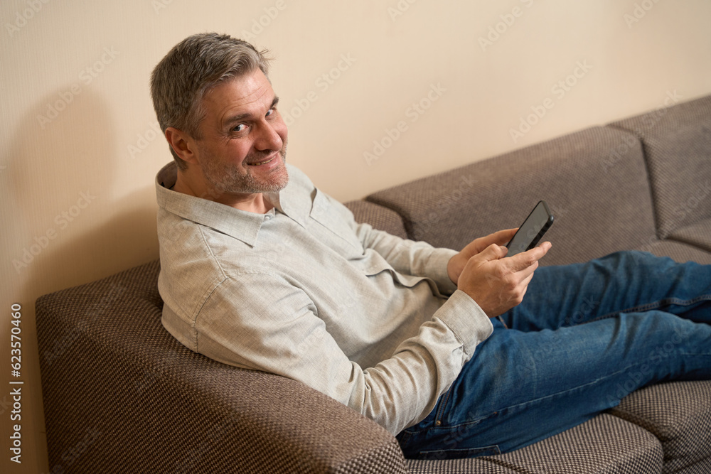 Smiling male holding phone and sitting on couch Smiling male holding phone and sitting on couch