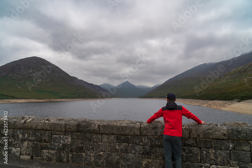 Misty morning in Mourne Mountains. Silent Valley water reservoir. Northen Ireland. Man in red hoodie standing on the edge of lake. Selective color