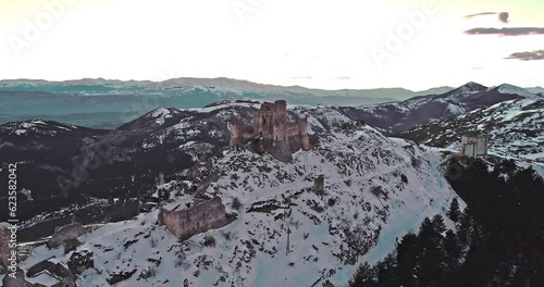 Aerial view of the Rocca di Calascio with snow and illuminated by the light of the sunset, behind the chain of the Gran Sasso and the Corno grande. Gran Sasso and Monti della Laga National Park, Abruz