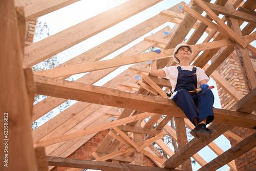 Bild auf Leinwand Woman roofer holding hammer, resting from ceiling work, sitting on roof girder
