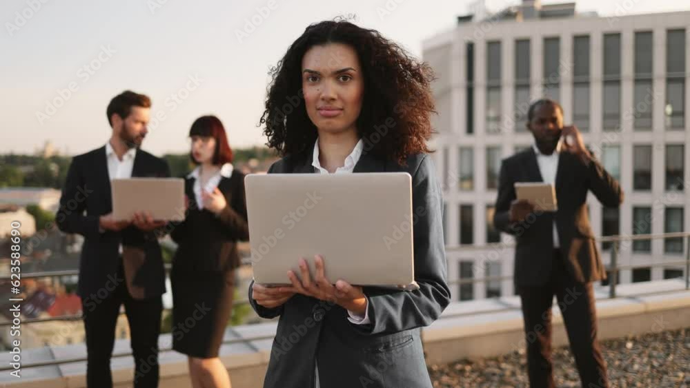 Portrait of pretty african american lady in business outfit posing on camera with laptop and smiling. Group of diverse entrepreneurs gathering on rooftop with gadgets to discuss new project plan. Portrait of pretty african american lady in business outfit posing on camera with laptop and smiling. Group of diverse entrepreneurs gathering on rooftop with gadgets to discuss new project plan.