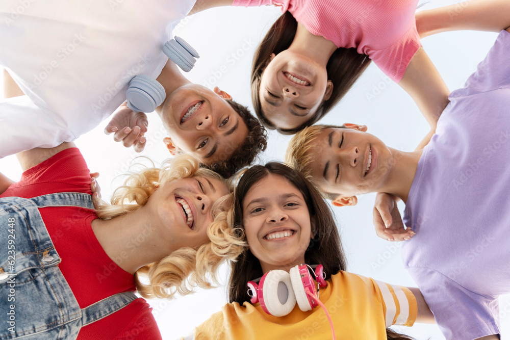 Fototapeta premium Group of happy multiracial teenagers, smiling friends wearing colorful t shirts embracing, looking at camera on street. Positive school boys and girls standing together. Vacation, summer, friendship