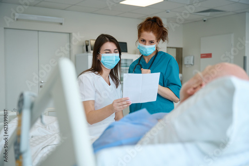 Female doctors with piece of paper visiting male patient in ward