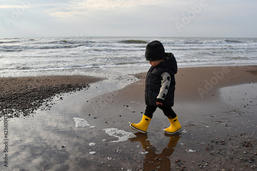 Little Toddler boy wearing yellow rubber boots walking down the sea in autumn season