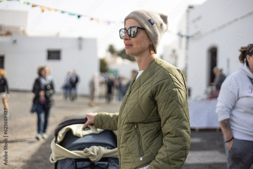 Portrait of young stylish woman wearing green padded jacket, hoodie, wool cap and sunglasses pershing stroller down cobled street of touristic town of Tequise, Lanyarote, Spain Portrait of young stylish woman wearing green padded jacket, hoodie, wool cap and sunglasses pershing stroller down cobled street of touristic town of Tequise, Lanyarote, Spain