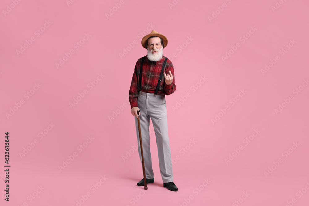 Fototapeta premium Senior man with walking cane showing his tongue and rock gesture on pink background