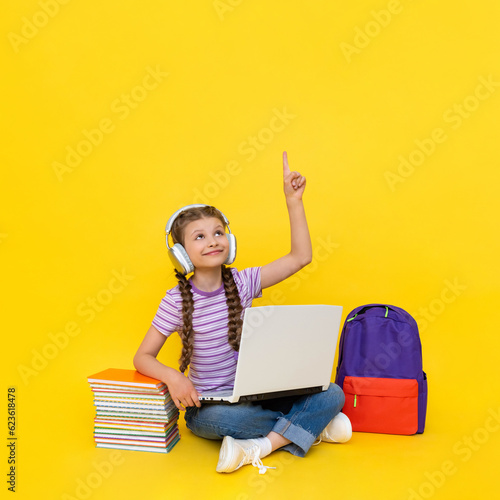 A young girl is sitting with a laptop in her hands and pointing up at your advertisement. A child with books, a backpack is engaged in an online school. Yellow isolated background.  Copy space.
