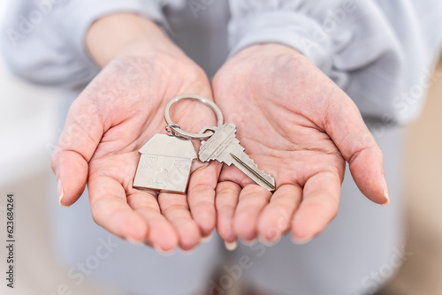 Female Hand Holding House Keys Inside Empty Room. Real estate agent handing over house keys in hand. Close-up view of keys from new home on cardboard box during relocation.