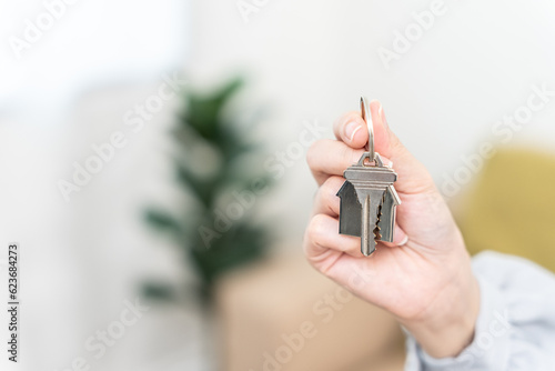 Female Hand Holding House Keys Inside Empty Room. Real estate agent handing over house keys in hand. Close-up view of keys from new home on cardboard box during relocation.