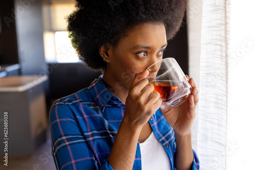 Thoughtful african american woman standing in kitchen drinking cup of tea, looking out of window photo