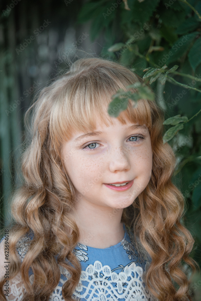 Fototapeta premium Portrait of a girl with long blonde hair on the street. A 9-11-year-old girl against a background of green plants, smiling and looking at the camera