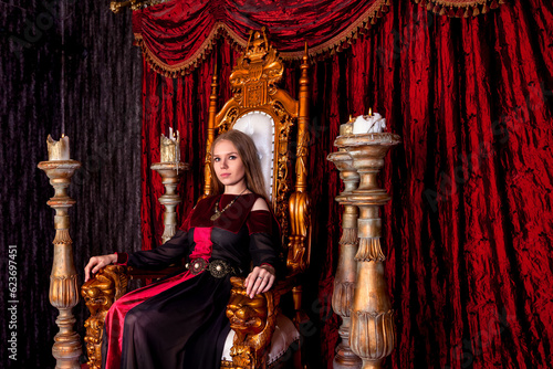 Lovely lady in an old style on antique throne fortress, looking at camera. Medieval queen in historical image posing sitting on golden throne in castle room. Concept of theatrical. Copy ad text space