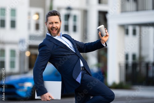 Excited businessman walking in city. Funny business man walk on city street. Business man walking outdoor, hold laptop take away coffee cup. Amazed business man in suit junp. Successful business.