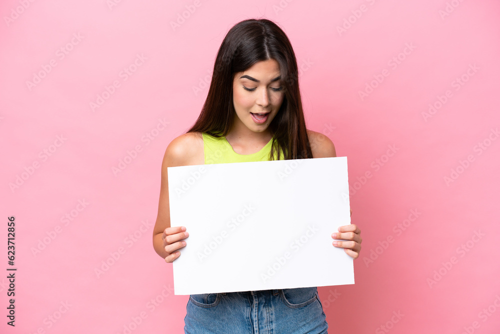 Young Brazilian woman isolated on pink background holding an empty placard and looking it Young Brazilian woman isolated on pink background holding an empty placard and looking it