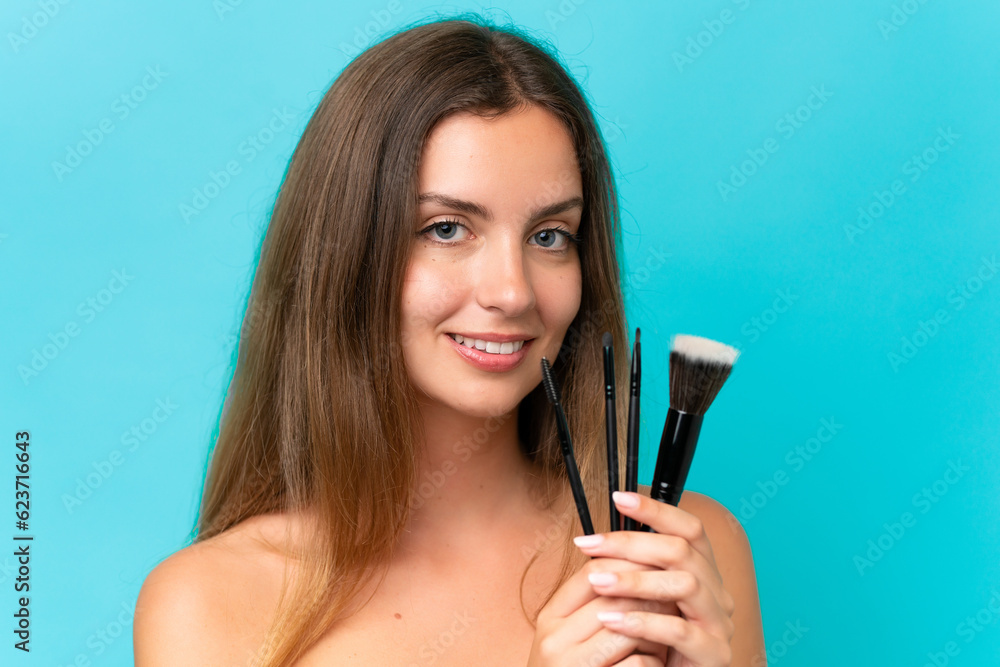 Young caucasian woman isolated on blue background holding makeup brush and whit happy expression Young caucasian woman isolated on blue background holding makeup brush and whit happy expression