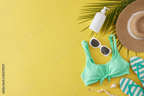 The idea of a summer beach relaxation. Top view shot of beach footwear, sunscreen bottle, swimsuit, hat, sunglasses, tropical leaves, seashells on yellow background with empty space for promo or text