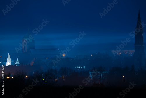 Zdjęcie przedstawia miasto nocą. Widać światła latarni ulicznych, oświetlone okna w budynkach i ciemne bryły wysokich budynków wież kościelnych.