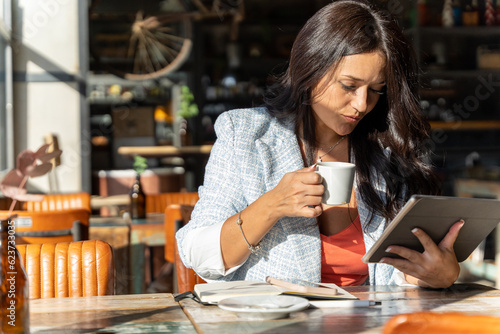 Woman looking at the digital tablet while drinking a cup of coffee in a coffee shop.