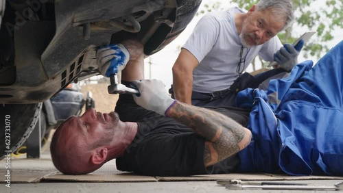 A car mechanic laying down on the ground using a wrench to screw in a bolt underneath a car while his supervisor watching behind