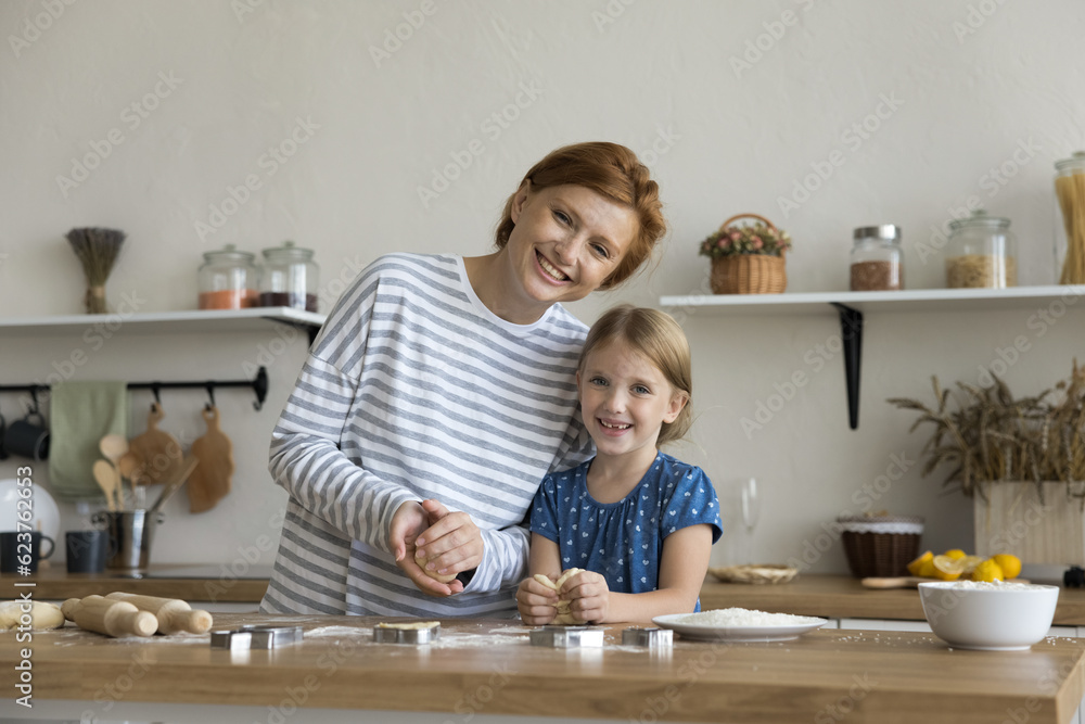 Happy sweet child girl and loving mom enjoying family culinary hobby together, baking cookies in home kitchen, holding raw dough over floury table, looking at camera with toothy smiles Happy sweet child girl and loving mom enjoying family culinary hobby together, baking cookies in home kitchen, holding raw dough over floury table, looking at camera with toothy smiles