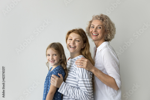 Happy girls and women of three generations posing at white wall background, hugging each other from behind, looking at camera with toothy smiles, laughing. Grandma, mom, kid side portrait