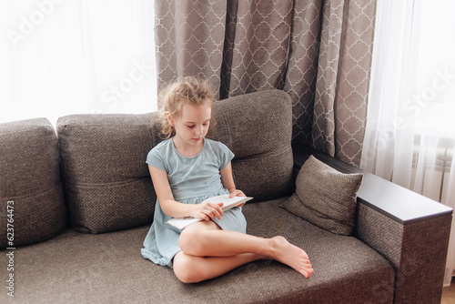A cute little girl in a blue dress is sitting on the couch reading a book