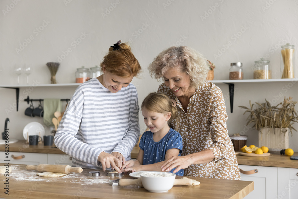 Cheerful girls and women of three family generations preparing sweet pastry food, baking biscuits together, cutting dough for cookies. Mom, grandma and child cooking in kitchen Cheerful girls and women of three family generations preparing sweet pastry food, baking biscuits together, cutting dough for cookies. Mom, grandma and child cooking in kitchen