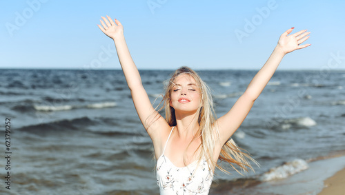 Happy blonde beautiful woman on the ocean beach standing in a white summer dress  raising hands