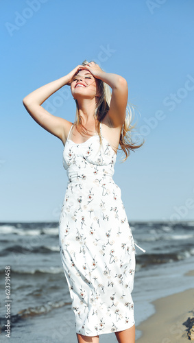 Happy blonde woman in free happiness bliss on ocean beach standing straight. Portrait of a female model in white summer dress enjoying nature during travel holidays vacation outdoors