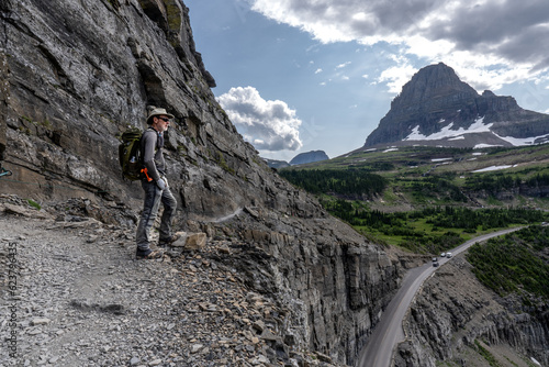 Matured Caucasian man, hiker, standing on a cliff watching a mountain scene while vehicles driving along a road below, Highline Trail, Going to the Sun Road, Glacier National Park, Montana
