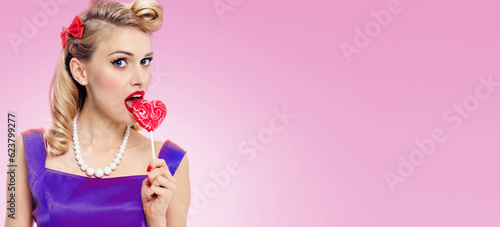 Woman eating heart shape lollipop dressed in pinup style dress in polka dot, over pink background. Caucasian blond model posing in retro fashion and vintage studio concept.