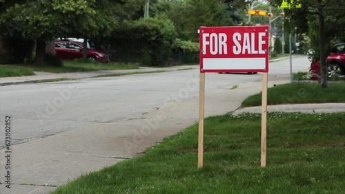generic for sale sign on front lawn of residential home in white writing on red background and wood posts with blurred long street behind, black car and white car passing - medium shot frame right photo